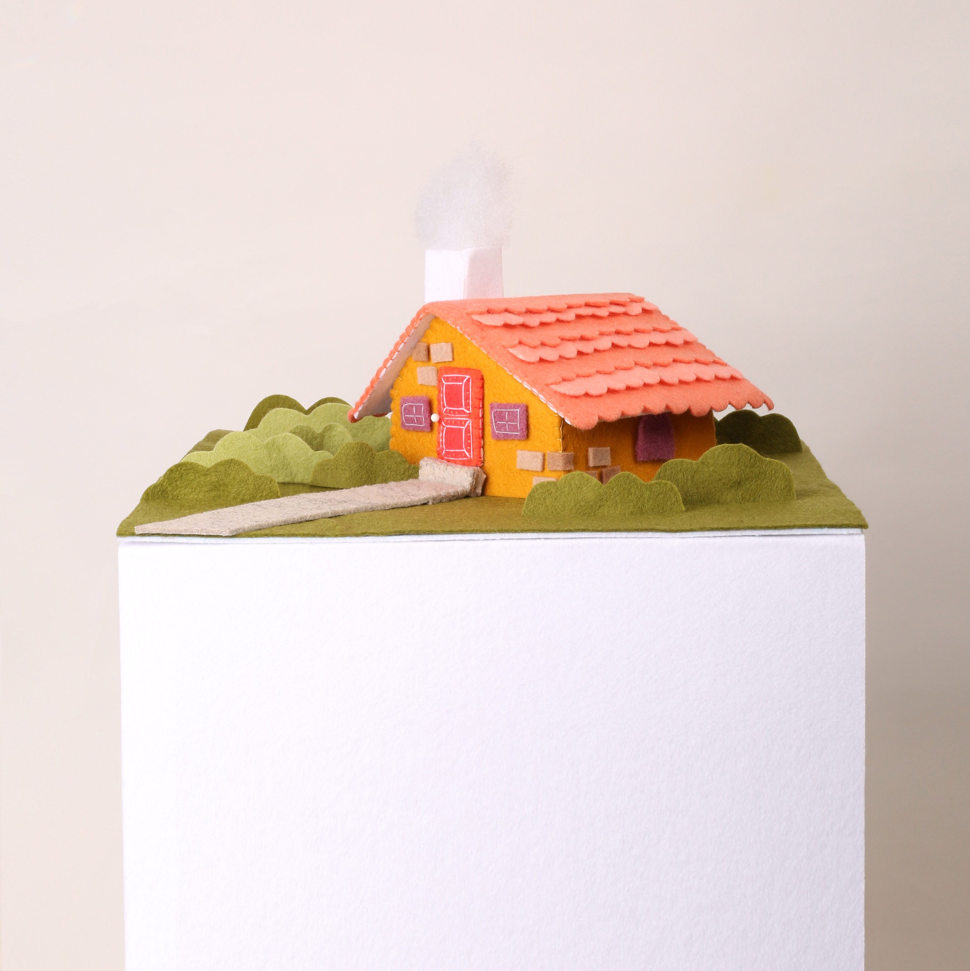 Tiny little felt house, with yellow walls and a red shingled roof, and a little puff of white smoke coming out of the chimney. A green grassy yard on little lumpy felt bushes. The house and garden is on a tall white plinth like it might be in a gallery.