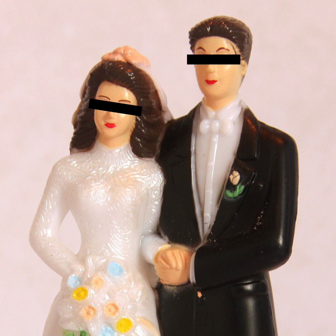 A wedding cake topper of a bride and groom with black bars superimposed over their eyes