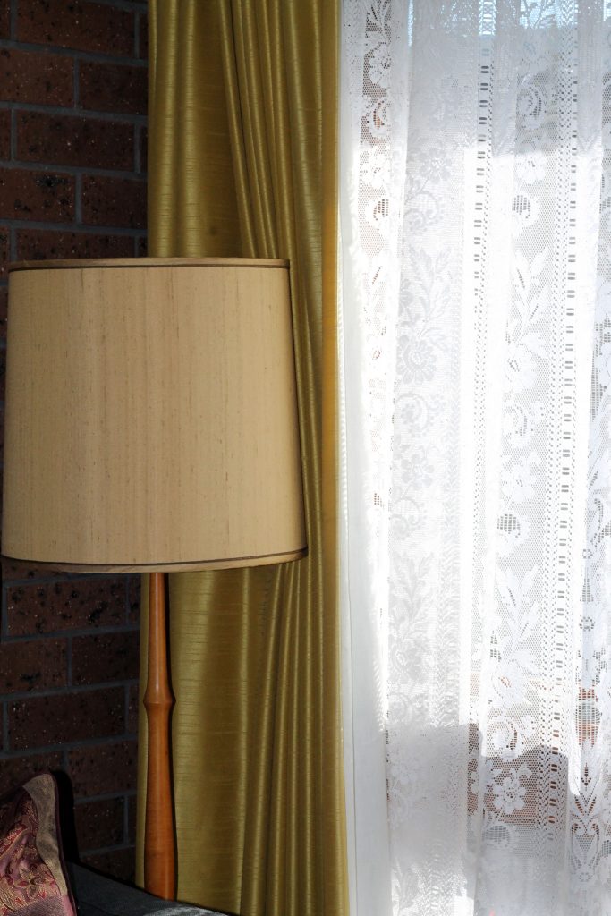 Photo of olive green velour pleated curtain opened to the left, opening upon a window covered by inner lace curtain. In front of green curtain is a white floor lamp with thin wooden stand.