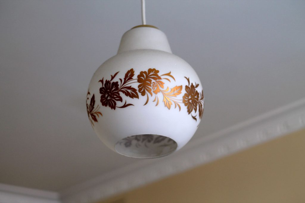 Photo of white light fixture with gold floral leaf design around its circumference. In background is the white ceiling meeting in the corner with relief skirting boards and one peach-coloured wall.