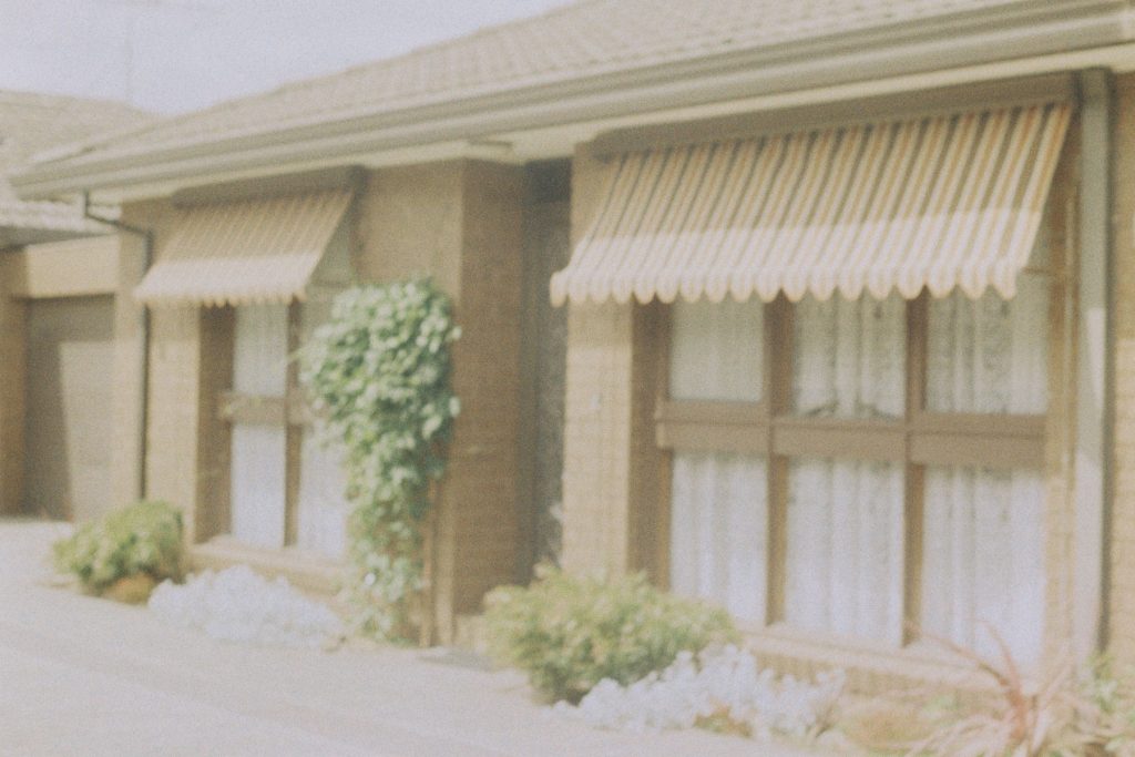 Over-exposed photograph of an old dark brick unit, one storey, with stripey 70s awnings over the floor-length windows, dotted by a few small shrubs.